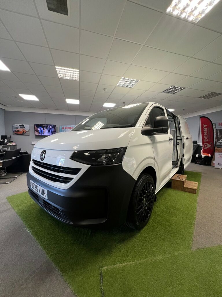 The 2025 ALL-NEW Volkswagen Transporter T7 150BHP Manual Commerce Plus Campervan in Clear White (Stock A1444) is showcased indoors on artificial grass, with its side door open and wooden steps leading inside. Office lights, posters, and a TV appear in the background.