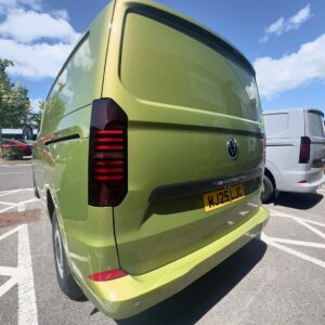 A Warm Green 2025 ALL-NEW Volkswagen Transporter T7 DSG Auto Commerce Plus Campervan (Deluxe SWB, Stock A1449) is parked in a sunny lot, with trees and a silver van in the background. The license plate reads WJ25 LJC.