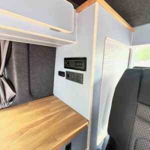 Close-up of the SWB T6.1 Volkswagen Transporter Highline Campervan (Candy White, 24 plate, A1447) interior with wooden countertop, grey cabinetry, electrical outlets, control panel, black seat, and a small window visible on the right.