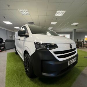 A 2025 ALL-NEW Volkswagen Transporter T7 150BHP Manual Commerce Plus Campervan in Clear White (Stock A1444) with black accents is parked on green carpet under fluorescent showroom lights, its license plate reading DE25 VUK.