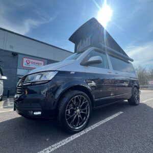A Reflex Silver LWB T6.1 Volkswagen Transporter Campervan (150BHP Manual, 24 Plate, A1455) with an elevated roof is parked outside a building displaying a red Denby sign on a sunny day.