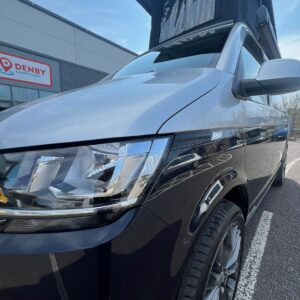 A Reflex Silver LWB T6.1 Volkswagen Transporter Campervan (150BHP Manual, 24 Plate, A1455) with its pop-up roof extended, parked outside the Denby Campervans building on a sunny day.
