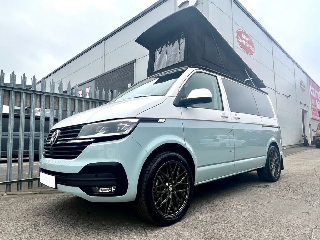 A SWB T6.1 Volkswagen Transporter Highline Campervan in Candy White (72 plate, A1451), featuring modern styling and black wheels, is parked outside a warehouse near a metal fence.