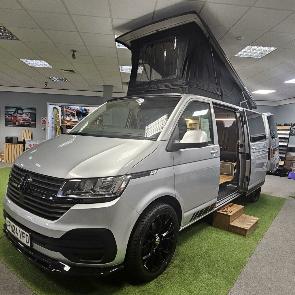 A LWB T6.1 Volkswagen Transporter Campervan (150BHP Manual, Reflex Silver, 24 Plate, A1457) with black wheels is parked on artificial grass in a showroom. The side door is open, showing a wooden interior and extended pop-up roof.