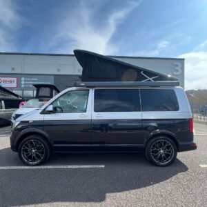 A LWB T6.1 Volkswagen Transporter Campervan 150BHP Manual in Reflex Silver (24 Plate, A1455) is parked in a lot on a sunny day, with a large industrial building and blue sky in the background.