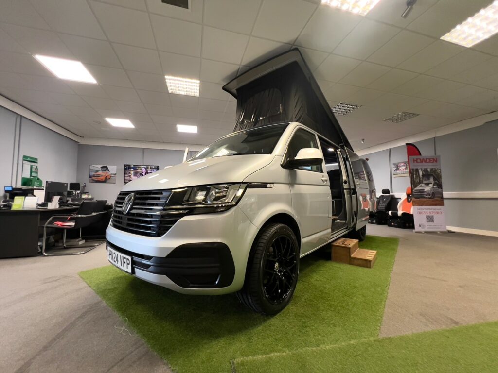 A Reflex Silver LWB Volkswagen T6.1 Transporter Campervan (150BHP Manual, 24 Plate, A1455) with a pop-up roof is displayed indoors on artificial grass, office desks and promotional banners in the background.