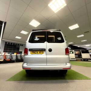 A LWB T6.1 Volkswagen Transporter Campervan, 150BHP manual in Reflex Silver (24 Plate, A1457), is displayed indoors on a green carpet, surrounded by office furniture and bright ceiling lights.