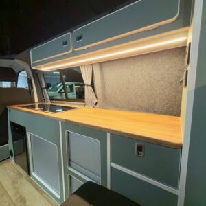 The LWB T6.1 Volkswagen Transporter Campervan 150BHP Manual (REFLEX SILVER – 24 Plate – A1457) features a modern interior with a wooden countertop, built-in cabinets, drawers, induction cooktop, under-cabinet lighting, and grey curtains.