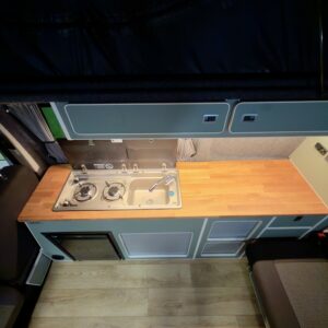 Overhead view of the LWB T6.1 Volkswagen Transporter Campervan (150BHP Manual, Reflex Silver, 24 Plate, A1457) kitchen featuring a wooden countertop, small sink, two-burner stove, cabinets, mini fridge, light wood floor & black upholstery.