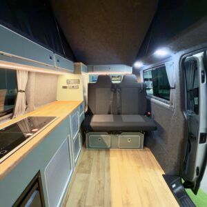 The LWB T6.1 Volkswagen Transporter Campervan 150BHP Manual (Reflex Silver, 24 Plate, A1457) features light wood flooring, a compact left-side kitchenette, foldable bench seat with storage, bright LED lighting, and an open side door.