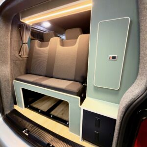 The LWB T6.1 Volkswagen Transporter Campervan (150BHP Manual, Reflex Silver, 24 Plate, A1457) has a cushioned bench seat, wood flooring, storage drawers, small cabinet, and warm LED strip lighting above in the rear.