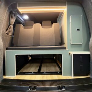 The image shows the interior of the LWB T6.1 Volkswagen Transporter Campervan (150BHP Manual, Reflex Silver, 24 Plate, A1457) with a gray and black bench seat, wooden floor, built-in storage, and a pull-out bed, seen from the open rear doors.