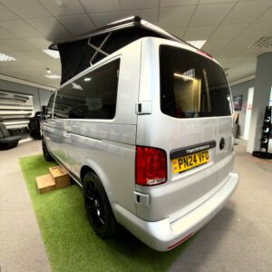 A LWB T6.1 Volkswagen Transporter Campervan (150BHP, Manual, Reflex Silver, 24 Plate, A1457) is parked indoors on artificial grass, with wooden steps to the side door and bright ceiling lights overhead.
