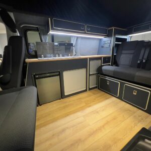 The LWB T6.1 Volkswagen Transporter Campervan (150BHP MANUAL, Reflex Silver, 24 Plate, A1455) features wood flooring, black cabinets, a kitchenette with sink and fridge, overhead storage, a black cushioned bench seat, and is well-lit and organized.
