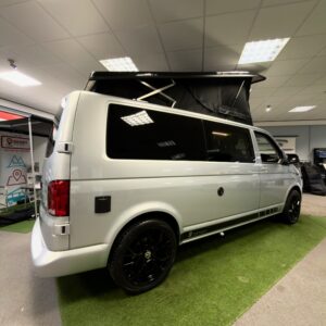 The LWB T6.1 Volkswagen Transporter Campervan 150BHP Manual in Reflex Silver (24 Plate, A1457) with black wheels and an elevated pop-top roof is displayed indoors on artificial grass, banners and other vehicles in the background.