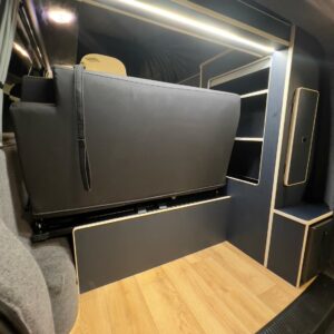The LWB T6.1 Volkswagen Transporter Campervan (150BHP Manual, Reflex Silver, 24 Plate A1455) features light wood flooring, dark cabinetry, open shelving, a foldable padded bench seat, and overhead LED strip lighting.
