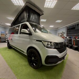 A Volkswagen Transporter Campervan (LWB T6.1, 150BHP Manual, Reflex Silver, 24 Plate, Ref: A1457) is displayed indoors on green carpet, surrounded by car seats and showroom lighting.