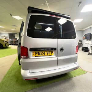 A LWB T6.1 Volkswagen Transporter Campervan 150BHP Manual in Reflex Silver (24 Plate, A1455) is parked indoors on green flooring with its rear facing the camera; office furniture and automotive displays are in the background.