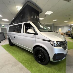The LWB T6.1 Volkswagen Transporter Campervan (150BHP Manual, Reflex Silver, 24 Plate, A1455) with a raised pop-up roof is showcased indoors on artificial grass, surrounded by office lights and another van in the background.