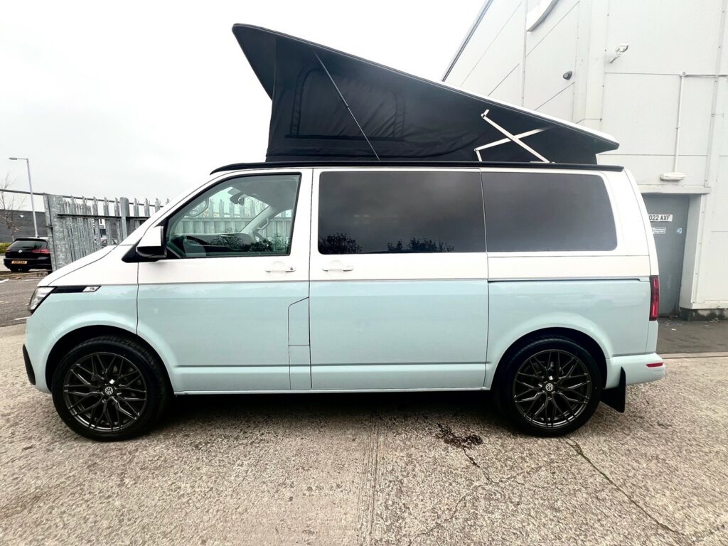 A Candy White VW T6.1 SWB Transporter Highline Campervan (72 plate, A1451) with a raised black pop-up roof, black wheels, and tinted windows is parked on concrete near a building and metal fence.