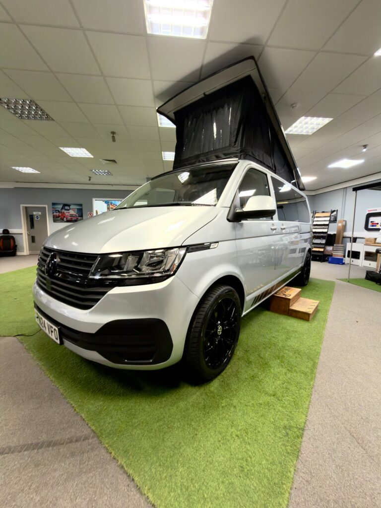 A LWB T6.1 Volkswagen Transporter Campervan (150BHP Manual, Reflex Silver, 24 Plate, A1457) with a raised pop-top roof is parked indoors on green artificial grass, with wooden steps by the sliding door under bright ceiling lights.