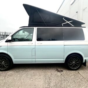 A Candy White VW T6.1 SWB Transporter Highline Campervan (72 plate, A1451) with a raised black pop-up roof, black wheels, and tinted windows is parked on concrete near a building and metal fence.