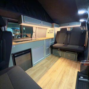 The LWB T6.1 Volkswagen Transporter Campervan (150BHP Manual, Reflex Silver, 24 Plate – A1457) features wood-look flooring, a compact kitchen with mini fridge, cabinets, cushioned bench seat, built-in lighting, and modern finishes.