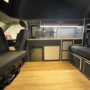 The LWB T6.1 Volkswagen Transporter Campervan (150BHP Manual, Reflex Silver, 24 Plate, A1455) features a modern interior with black and wood accents, built-in seating, storage cabinets, a kitchenette with mini fridge, and wood-style flooring.
