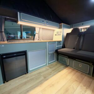 The LWB - T6.1 Volkswagen Transporter Campervan (150BHP MANUAL, Reflex Silver, 24 Plate, A1457) features wood flooring, gray cabinets, a compact fridge, overhead storage, curtained window, cushioned bench with seatbelts, and soft lighting for a clean look.