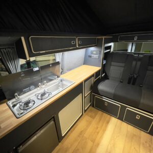 The LWB T6.1 Volkswagen Transporter Campervan (150BHP MANUAL, Reflex Silver, 24 Plate, A1455) features a modern interior with compact kitchen: two-burner stove, sink, wood countertop, black cabinets, cushioned bench seat, and light wood flooring.