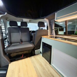 The LWB T6.1 Volkswagen Transporter Campervan (150BHP Manual, Reflex Silver, 24 Plate – A1457) features wood flooring, a gray bench seat, a compact kitchenette with cabinets and mini fridge, plus the driver’s area visible in the background.