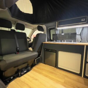 Interior of the LWB T6.1 Volkswagen Transporter Campervan (150BHP Manual, Reflex Silver, 24 Plate – A1455) featuring a black bench seat, kitchenette with sink and stove, mini fridge, overhead cabinets, wood-style flooring, and raised roof for space.