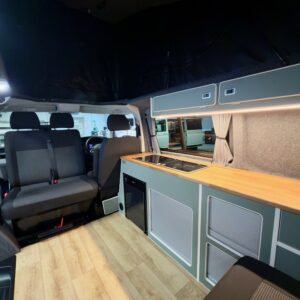 The LWB T6.1 Volkswagen Transporter Campervan (150BHP Manual, Reflex Silver, 24 Plate, A1457) features a modern interior with wooden flooring, kitchenette (countertop, sink, storage), two gray seats, and large windows.