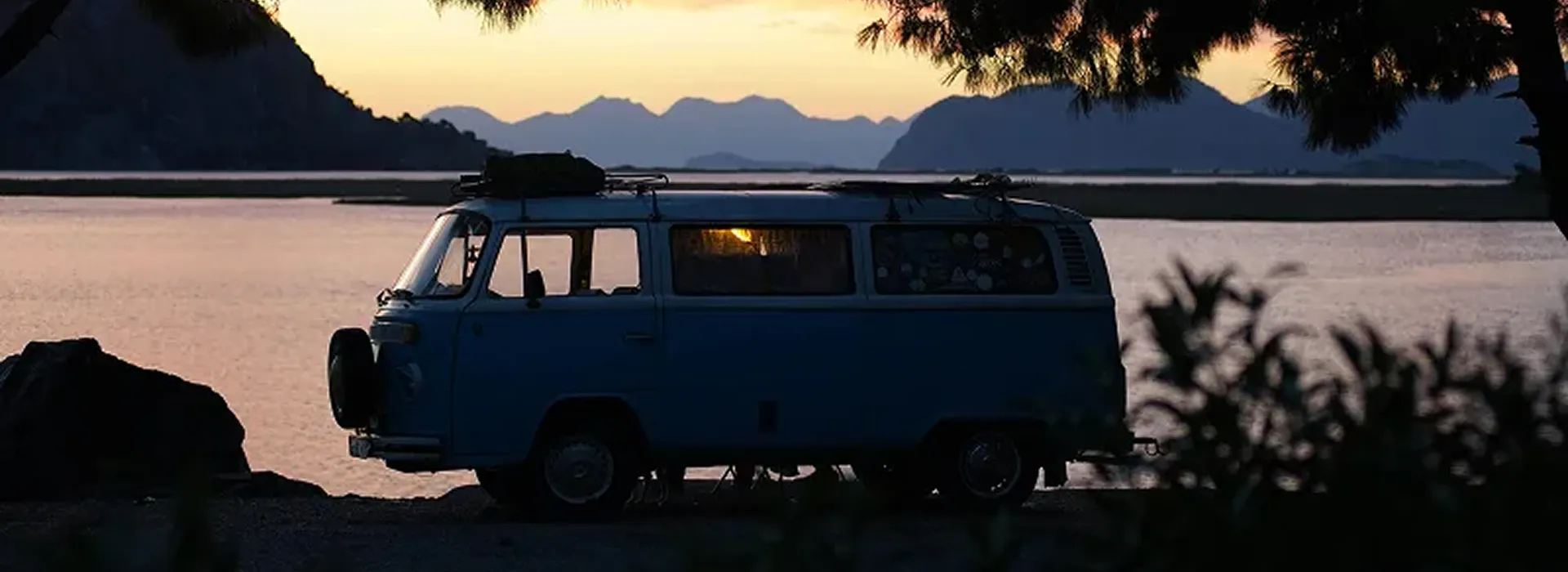 A blue van is parked by a calm lake at sunset, with silhouetted mountains and trees in the background and a warm light glowing inside the van.