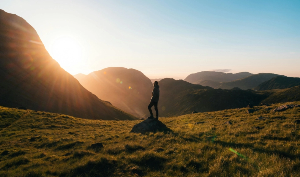 A person stands on a rock in a grassy mountain valley at sunrise or sunset, with sunlight streaming over distant mountains and casting long shadows across the landscape.