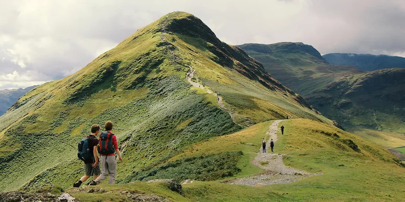 Hikers walk along a grassy, winding trail leading up a green hill under a partly cloudy sky, with mountains in the background.
