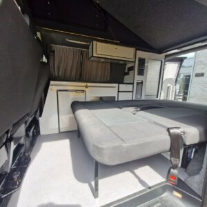 The SWB T6 Volkswagen Transporter Highline Campervan (Deep Black, 17 Plate, A1466) features a folded-down bed, black and white cabinetry, small fridge, window curtains, and sunlight streaming through the side door.