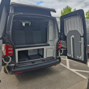 The image shows the rear of a SWB - T6 Volkswagen Transporter Highline Campervan in Deep Black (17 Plate, A1466), with its doors open to reveal a foldable bench seat, storage compartments, and a curtained window. The van is parked on a cloudy day.