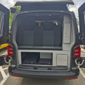 The SWB T6 Volkswagen Transporter Highline Campervan (Deep Black, 17 Plate, A1466) is parked outdoors with its rear doors open, revealing a grey bench seat, storage cabinets, and drawers in an organized interior.