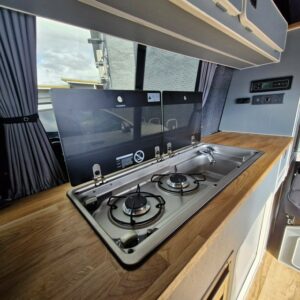 The SWB T6.1 Volkswagen Transporter Highline Campervan (Automatic, Ravenna Blue, 24 plate, A1471) features a compact kitchen with a wooden countertop, two-burner gas stove and sink under a glass lid; curtains and control panels visible behind.