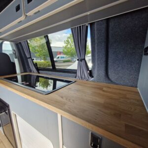 The SWB T6.1 Volkswagen Transporter Highline Campervan (Automatic, Ravenna Blue, 24 plate A1471) features a modern interior with a wooden countertop, black glass stovetop, grey cabinets, grey curtains, and scenic views outside.