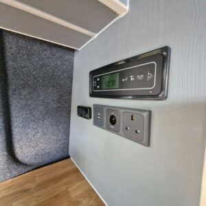 A close-up of the SWB - T6.1 Volkswagen Transporter Highline Campervan’s wall panel on a gray wall above a wood floor, showing a digital display, control buttons, USB port, European and UK power outlets.