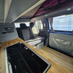 The SWB T6.1 Volkswagen Transporter Highline Campervan in Ravenna Blue (24 plate, A1471) features a wooden floor, black glass cooktop kitchenette, gray belted seats, curtained windows, and storage under seats and along the walls.