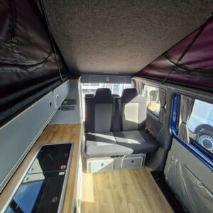 The SWB T6.1 Volkswagen Transporter Highline Campervan (Automatic, Ravenna Blue, 24 plate, A1471) features wood flooring, a left-side kitchenette with stove and sink, overhead cabinets, and a gray bench seat with belts and storage by the window.