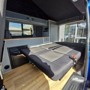 The image shows the interior of an SWB T6.1 Volkswagen Transporter Highline Campervan (Automatic, Ravenna Blue, 24 plate, A1471) with a folded-down bed, grey upholstery, wood-effect flooring, storage cabinets, and a small refrigerator lit by natural light.