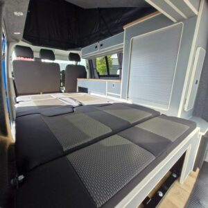 Interior of the SWB T6.1 Volkswagen Transporter Highline Campervan (Automatic, Ravenna Blue, 24 plate A1471) with grey seats folded into a bed, storage, curtained windows, modern light cabinetry and natural light flooding in.
