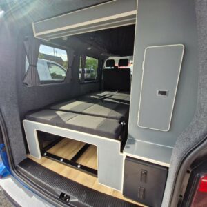The SWB T6.1 Volkswagen Transporter Highline Campervan (Automatic, Ravenna Blue, 24 plate, A1471) features a folded-out bed, grey fabric walls, curtained windows, built-in storage, and visible front seats through the open space.