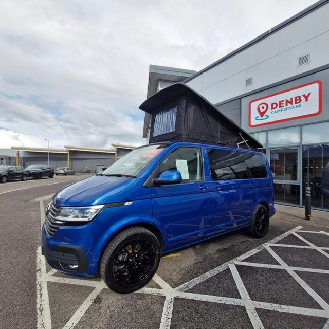 A Ravenna Blue SWB T6.1 Volkswagen Transporter Highline Campervan (24 plate, Automatic, A1471) is parked outside a Denby Campervans showroom with large glass windows and a red and white Denby sign on a cloudy day.