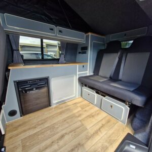 The SWB T6.1 Volkswagen Transporter Highline Campervan (Automatic, Ravenna Blue, 24 plate, A1471) features wood-look flooring, built-in storage cabinets, a small fridge, curtained windows, and a cushioned bench seat with seatbelts in a compact modern interior.