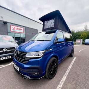 A Ravenna Blue SWB T6.1 Volkswagen Transporter Highline Campervan (Automatic, 24 plate, A1471) is parked outside a Denby building next to another Volkswagen. The sky is partly cloudy with trees in the background.